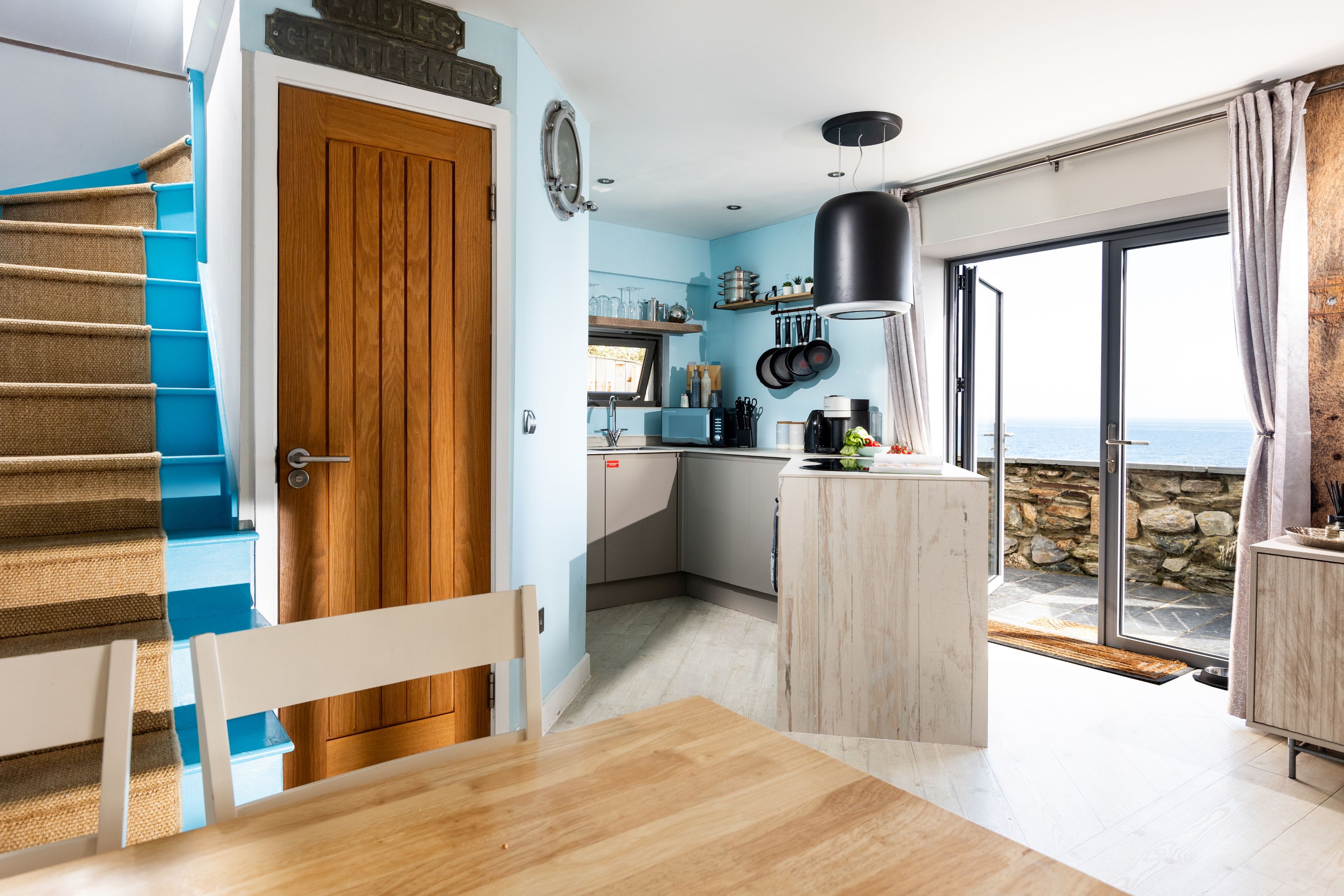 A modern kitchen with a wooden door, a dining area with a table, and a view of the sea through a glass door.