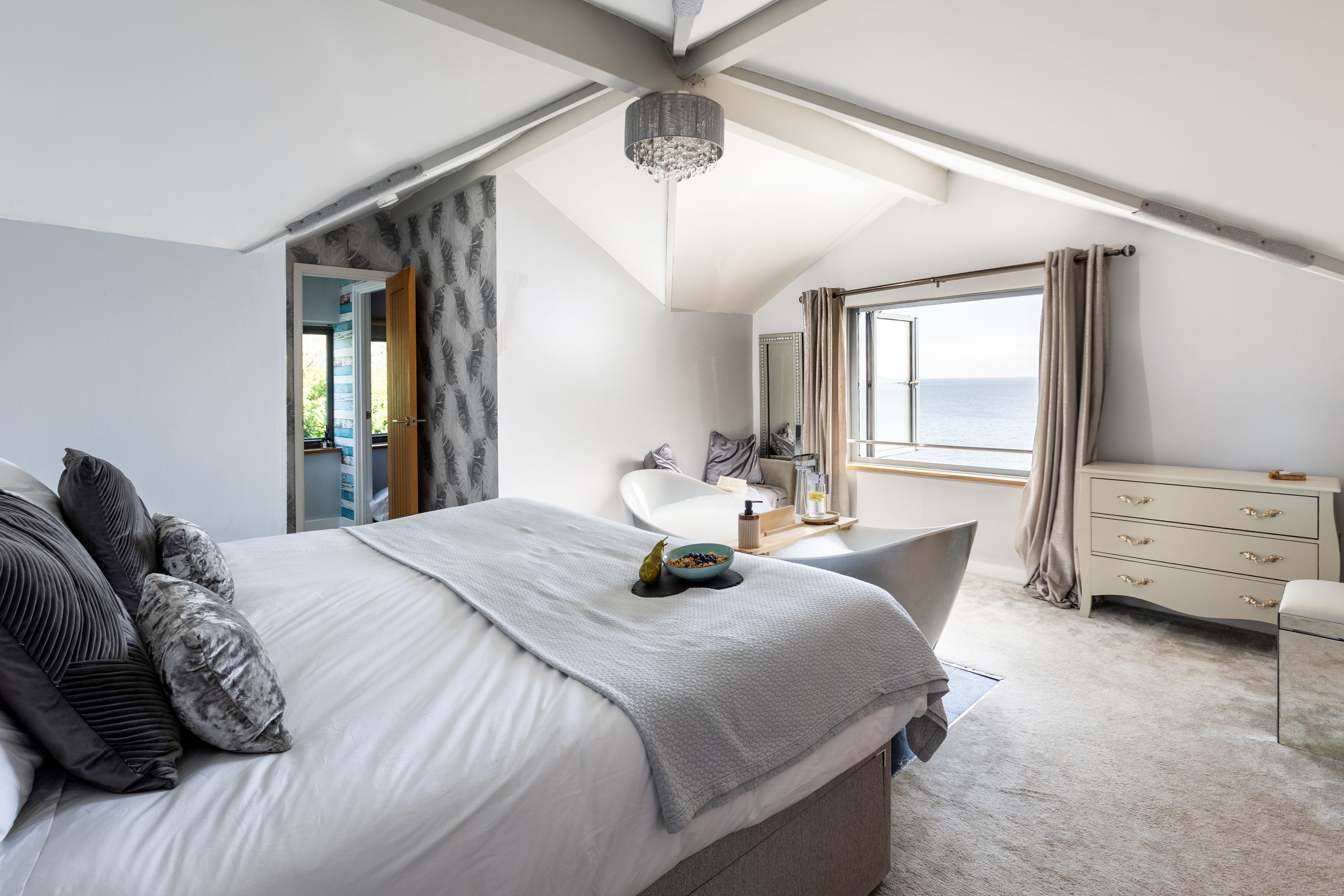 A bedroom with a large bed, a dresser, a vanity, and a view of the sea.