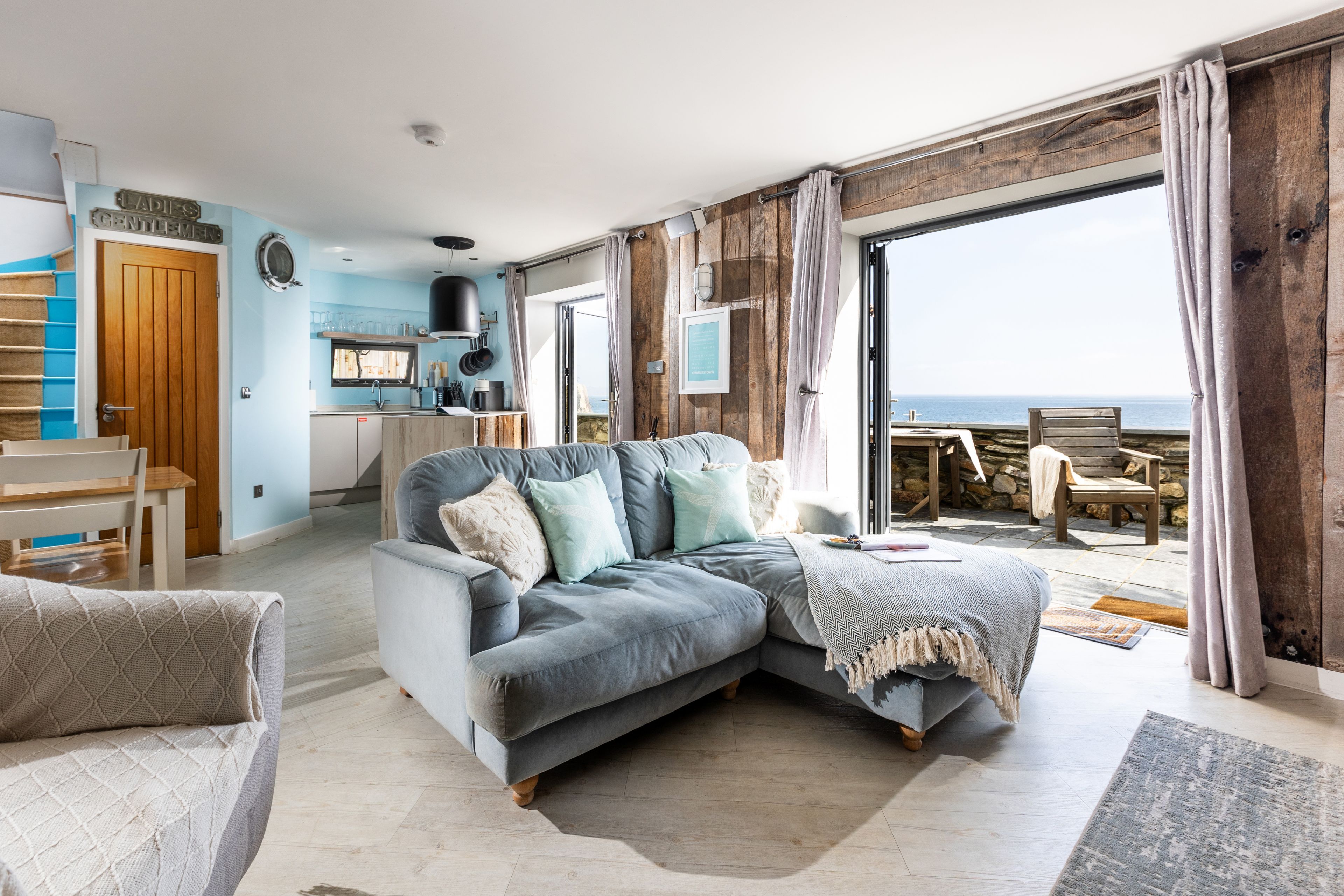 A modern living room with a grey sectional sofa, a dining area with a wooden table and chairs, and a kitchen with a window overlooking the sea.