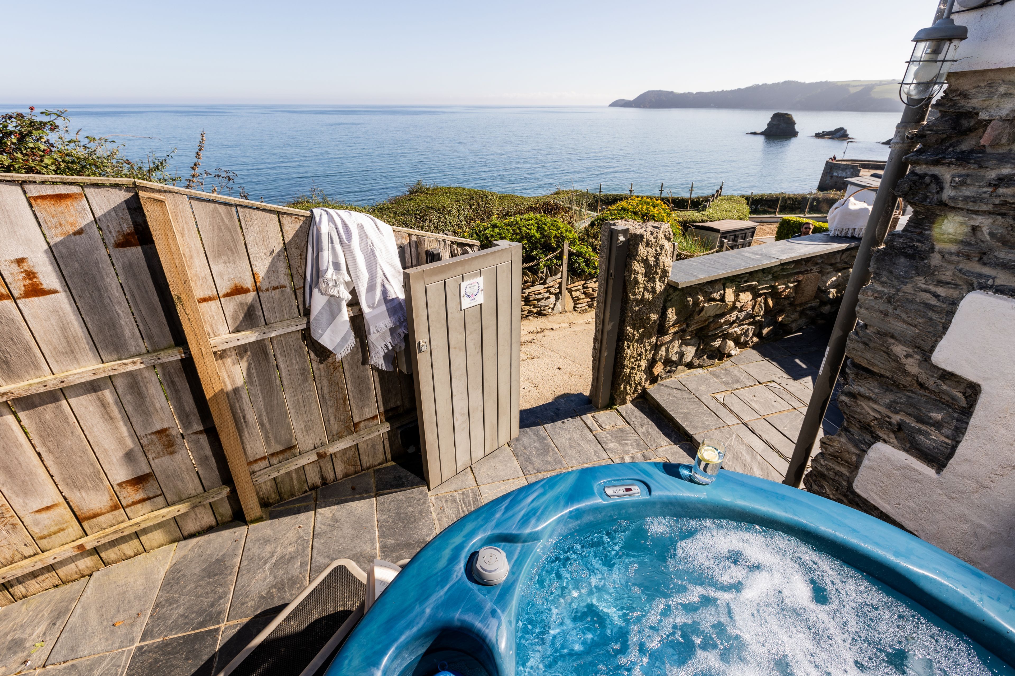 A wooden deck with a hot tub, a stone wall, and a view of the sea.