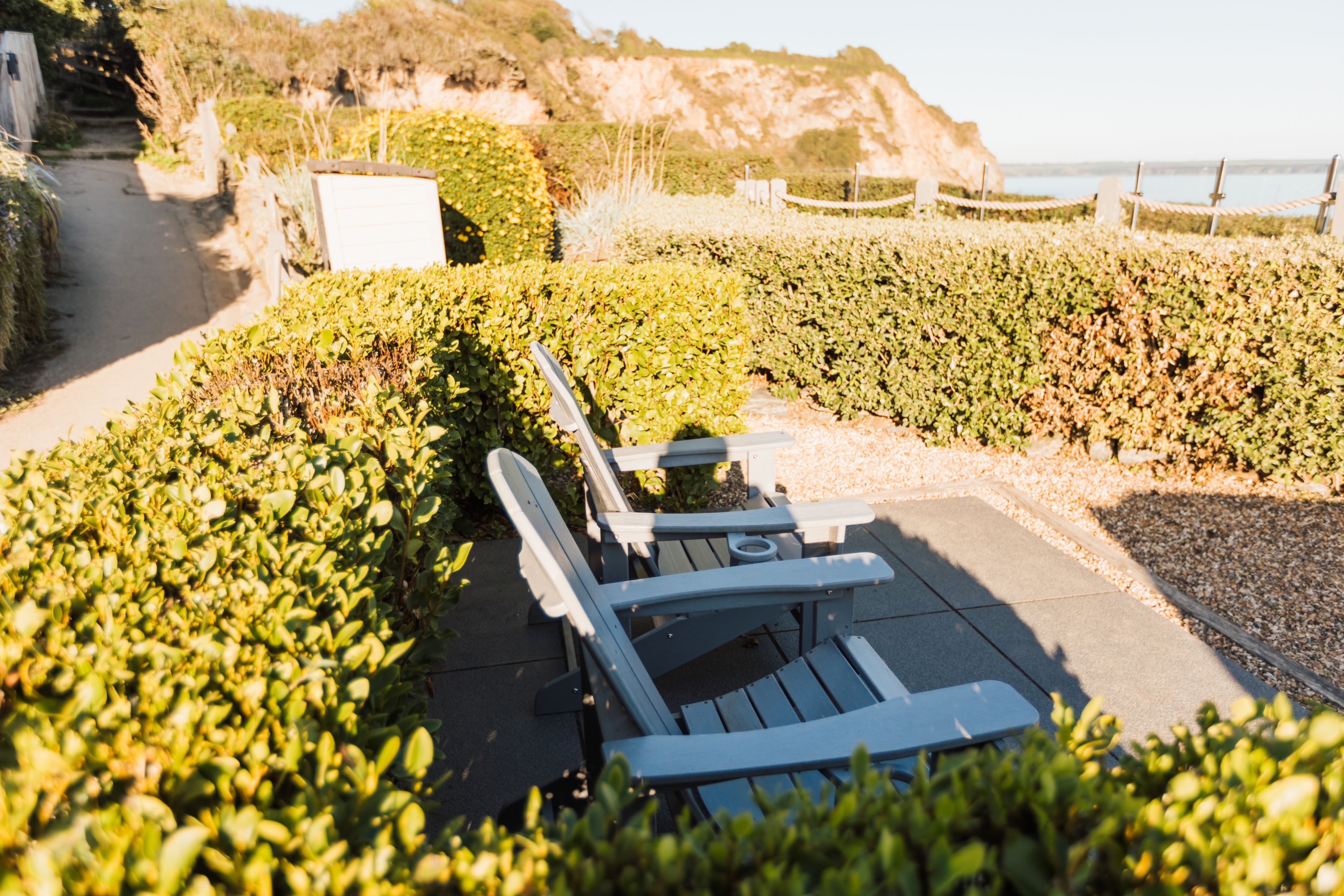 A paved outdoor seating area with a table and chairs, surrounded by neatly trimmed hedges and a gravel path.
