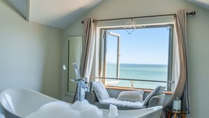 A modern bathroom with a freestanding bathtub, a seating area with a view of the sea, and a small table with a candle. - As seen on TV! Luxurious Beach Front Cottage with Hot Tub, Log Burner & Sea Views (Charlestown)
