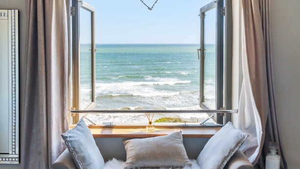 A room with a view of the ocean through an open window, a cushioned bench with pillows, and a small table with a bottle. - As seen on TV! Luxurious Beach Front Cottage with Hot Tub, Log Burner & Sea Views (Charlestown)
