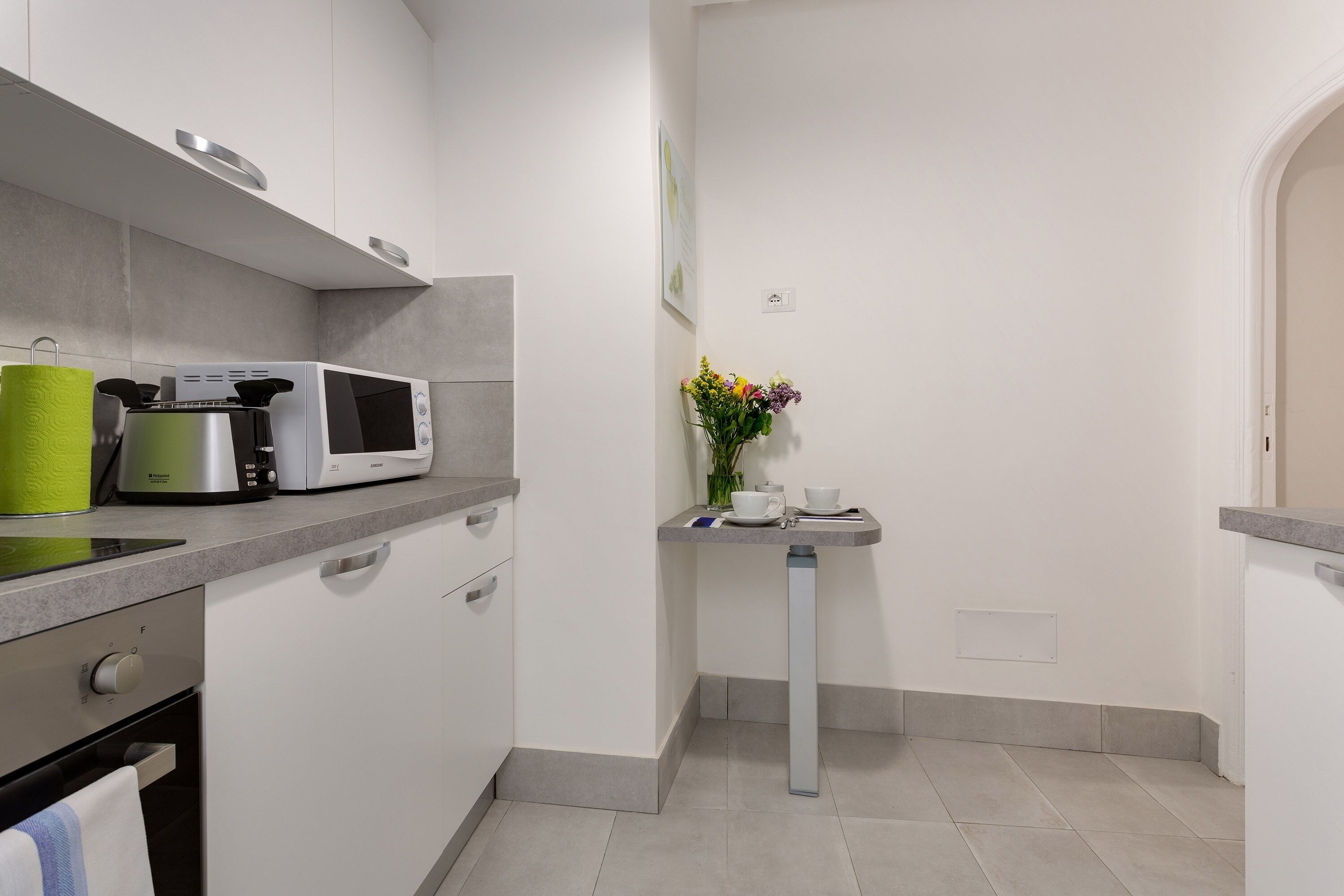 A modern kitchen with white cabinets, a microwave, a kettle, and a small table with a vase of flowers.