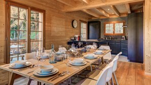 Un intérieur en bois avec une table à manger dressée pour six personnes, une cuisine aux meubles sombres et de grandes fenêtres.