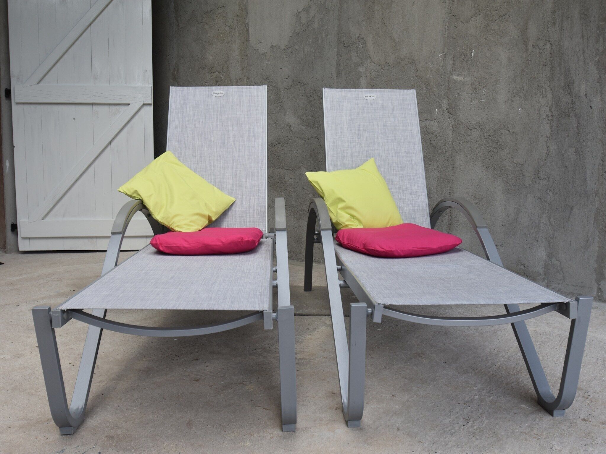 Two grey lounge chairs with metal frames and cushions, positioned against a concrete wall with a white door in the background.