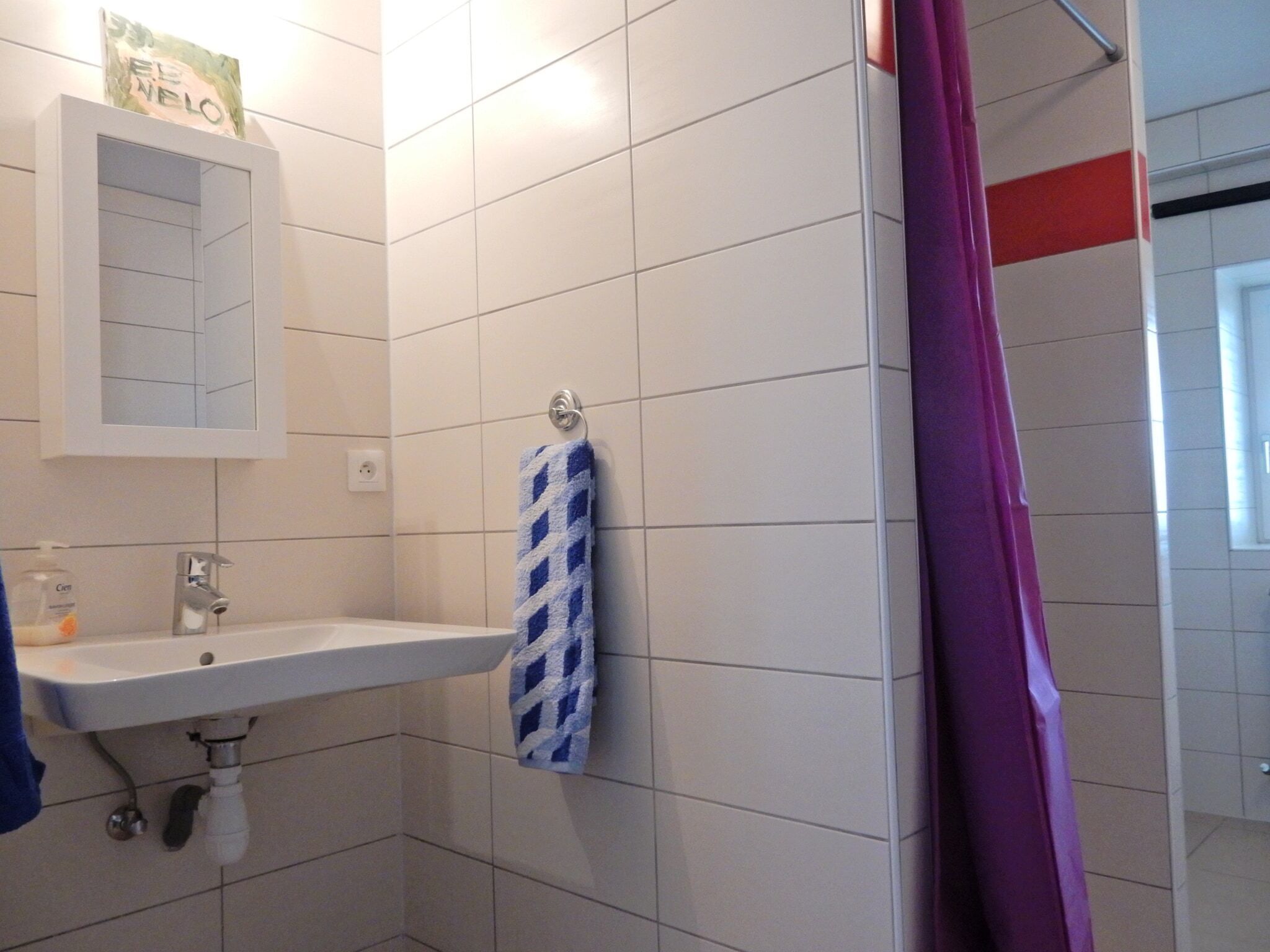 Un baño con paredes de azulejos blancos, un lavamanos blanco, un espejo y una toalla azul colgada en la pared.