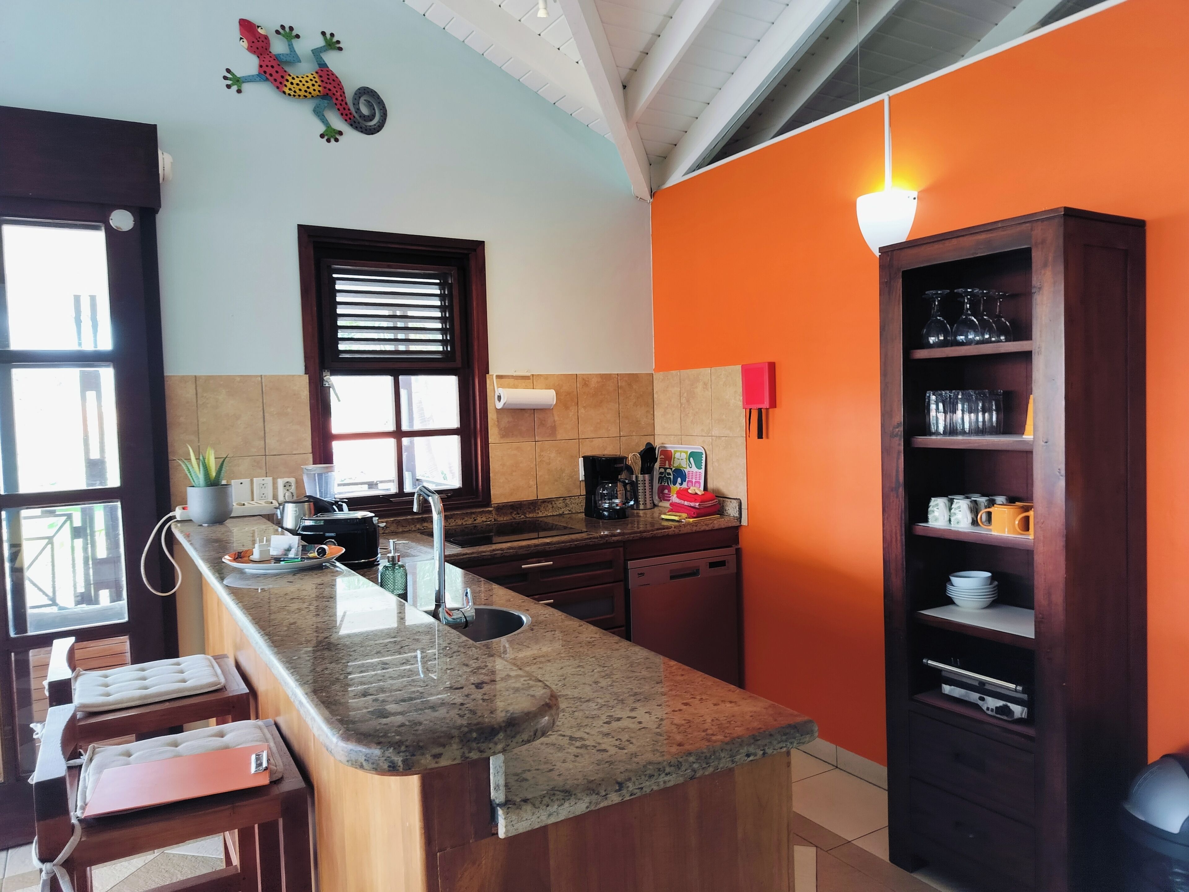 A kitchen with a granite countertop, a sink, a window with blinds, a red wall, and a shelf with glassware.
