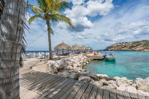 A wooden deck leading to a beach with thatched umbrellas, lounge chairs, and a rocky shoreline.