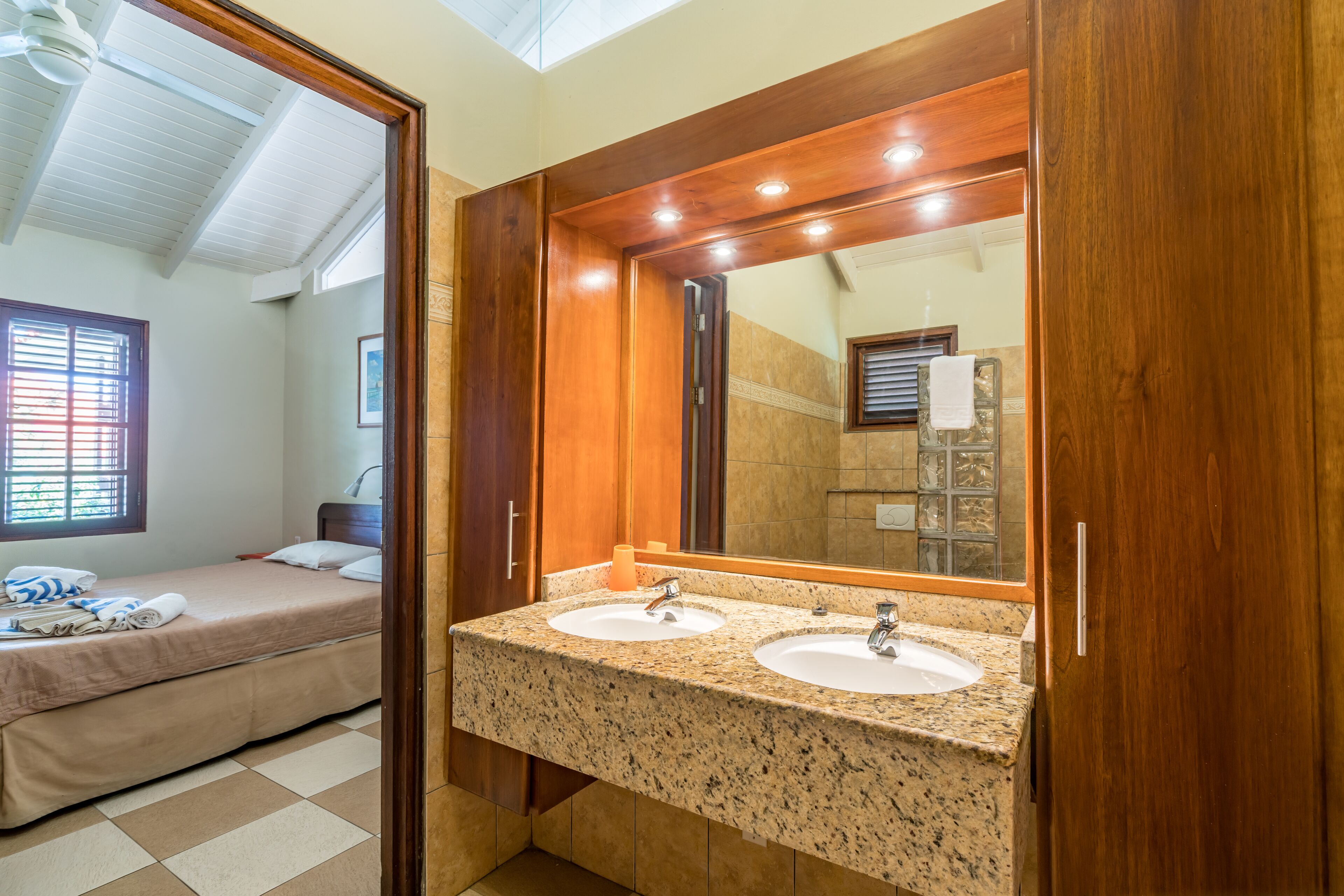 A hotel bathroom with two sinks, a large mirror, and a bed in the adjacent room.