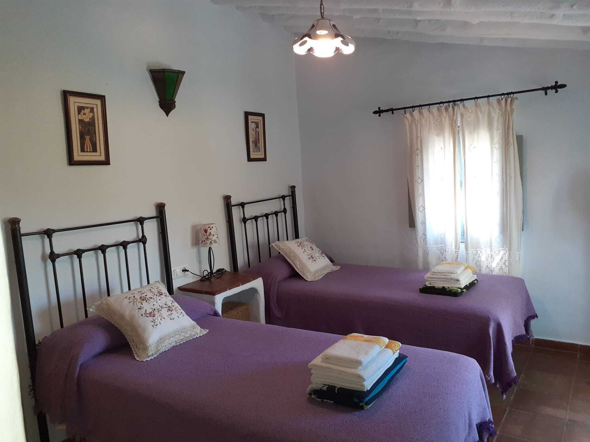 Ein Zimmer mit zwei Betten, jeweils mit violetter Bettdecke und Kissen. Dazwischen steht ein kleiner Tisch mit einer Lampe.
