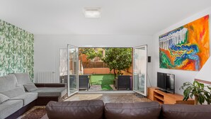Un salon moderne avec un canapé gris, un divan en cuir marron, un téléviseur posé sur un meuble en bois et un tableau abstrait aux couleurs vives accroché au mur.