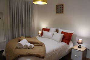 Un lit bien fait, avec des oreillers blancs et rouges, une couverture marron et des serviettes enroulées. On y trouve une table de chevet avec une lampe et un petit tiroir.