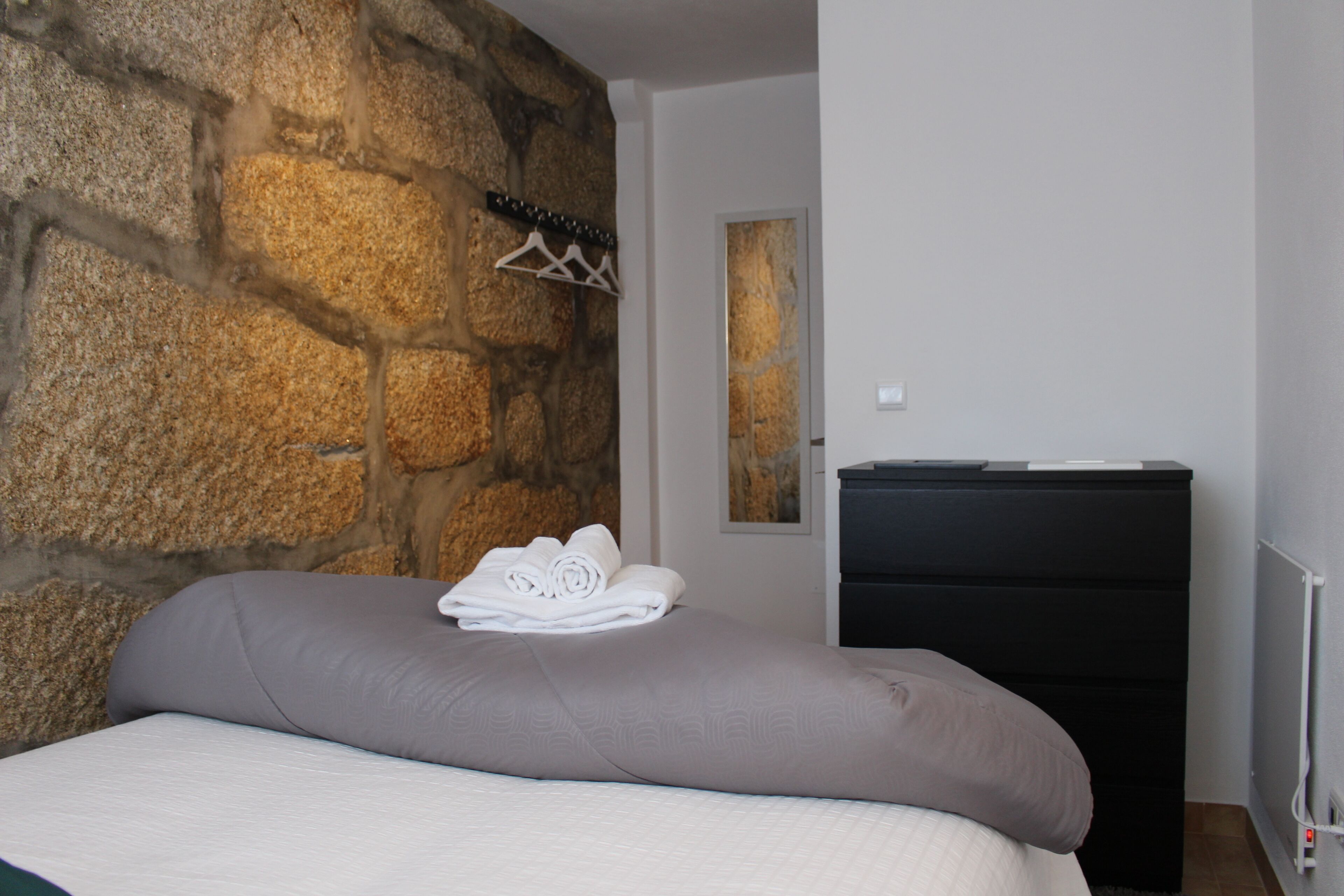 A bedroom with a stone wall, a bed with rolled towels, a black dresser, and a white hanging rack.