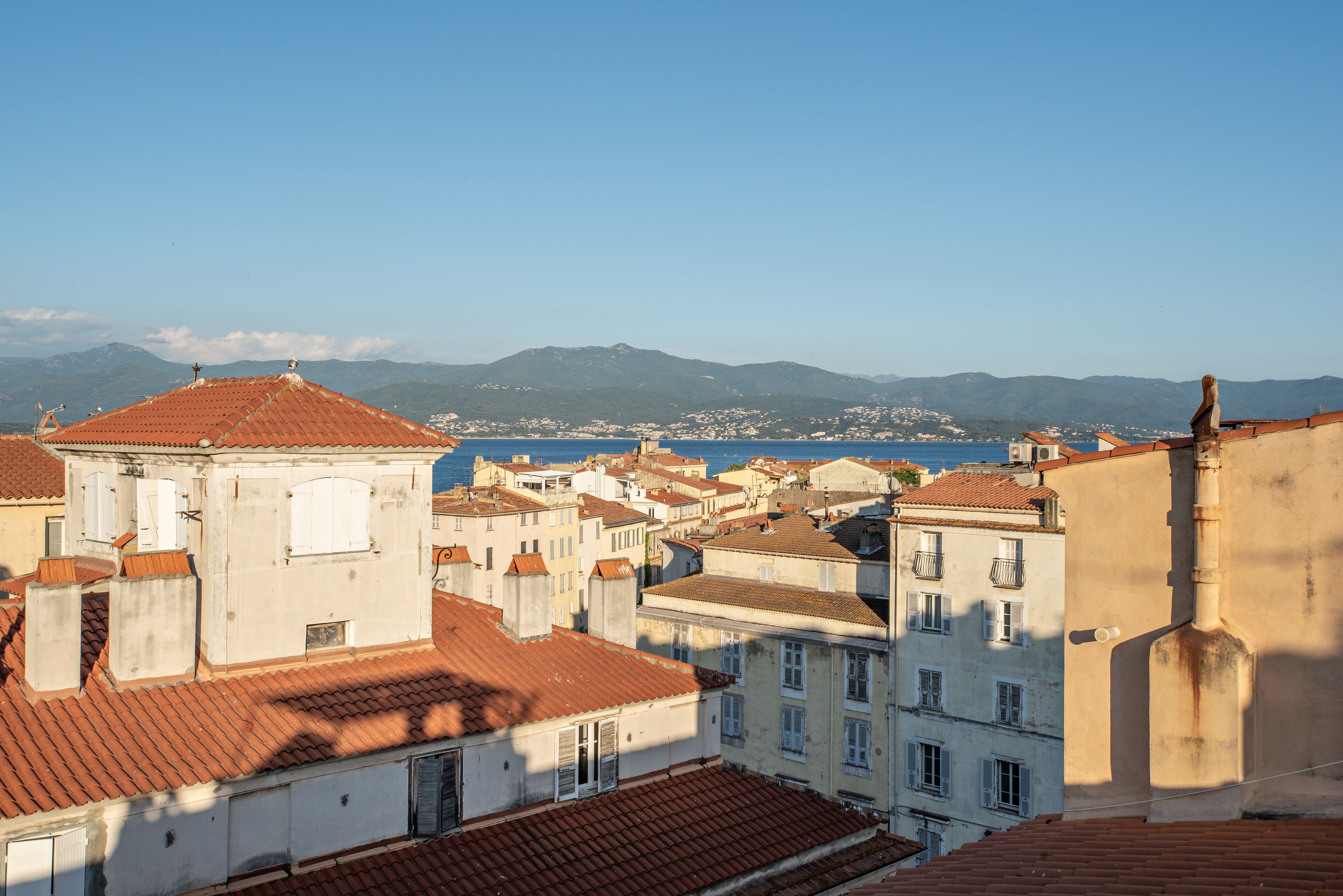 A coastal cityscape with terracotta-roofed buildings, a clear blue sky, and distant mountains.