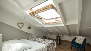 Um quarto com claraboia, uma cama com roupa de cama branca, uma cadeira azul e uma pequena mesa redonda.