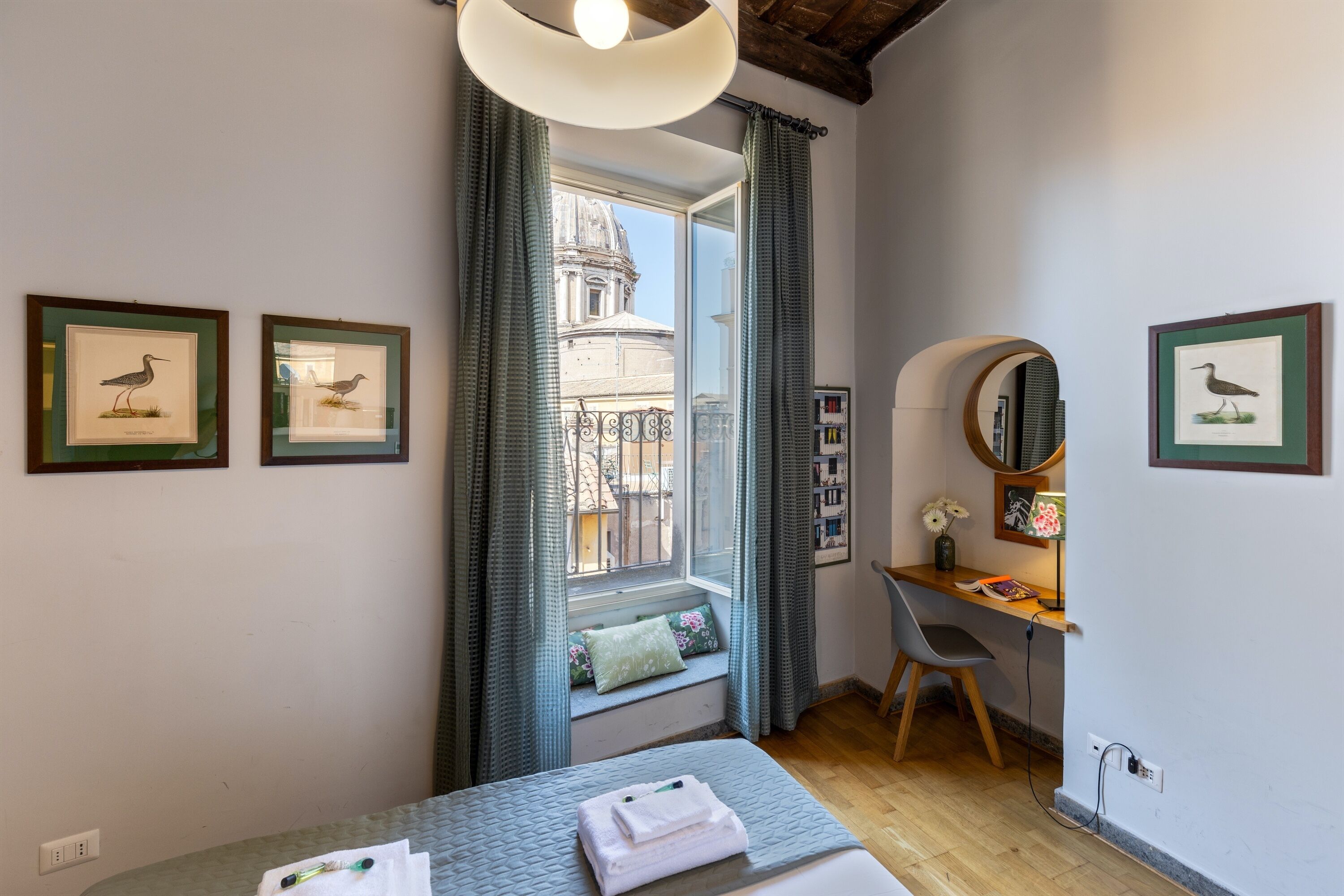 A bedroom with a bed, a desk, and a window with curtains. There are framed pictures on the wall.