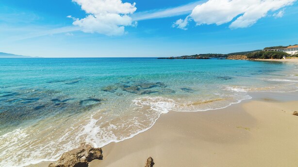 A beach with clear turquoise water, sandy shore, and a distant building on a hill.