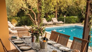 An outdoor dining area with a pool view, wooden chairs, a table set with plates, glasses, and a bottle of champagne.
