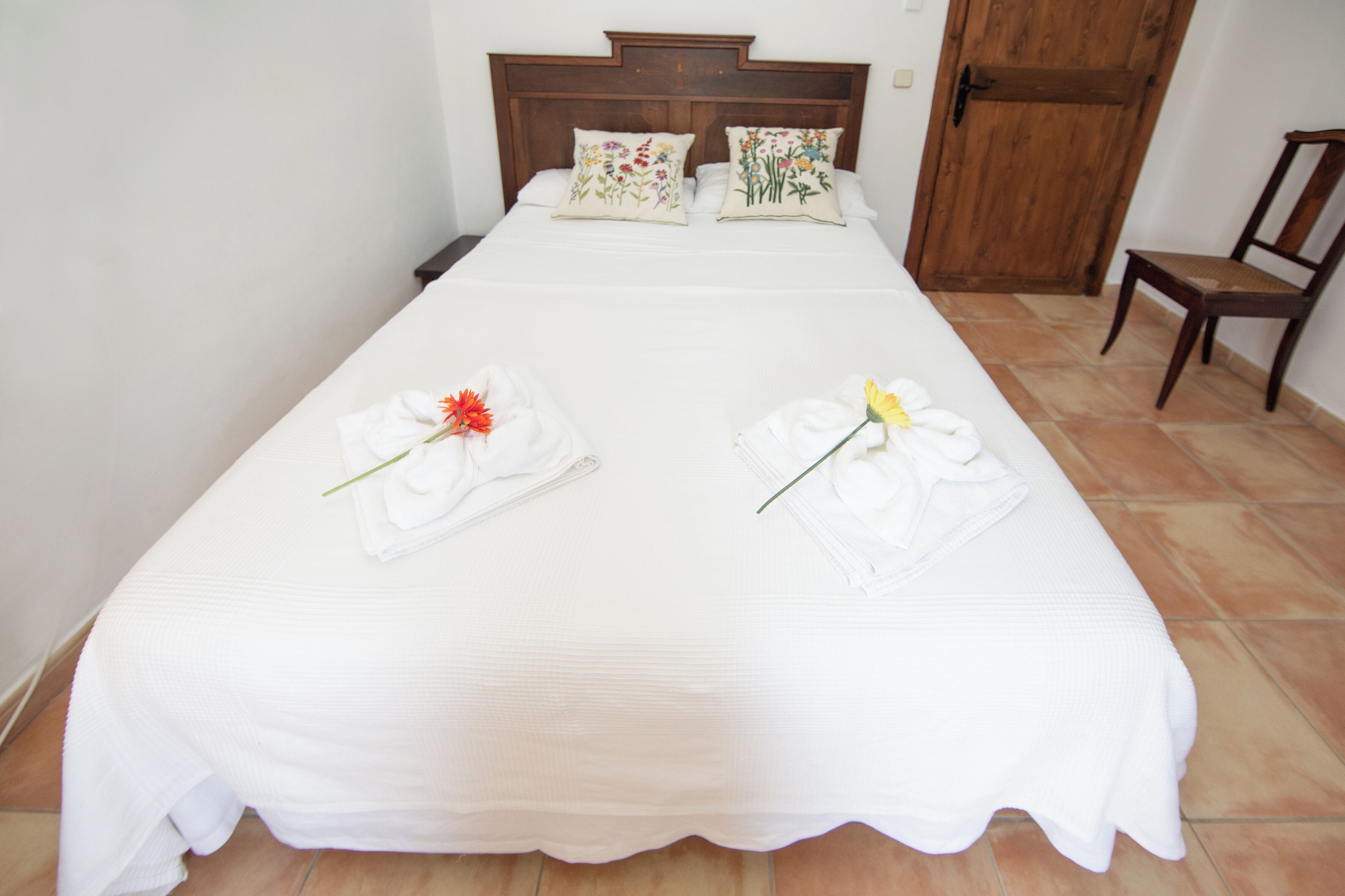 Cama bem arrumada com lençóis brancos e duas toalhas com flores, uma cabeceira de madeira e uma porta de madeira ao fundo.