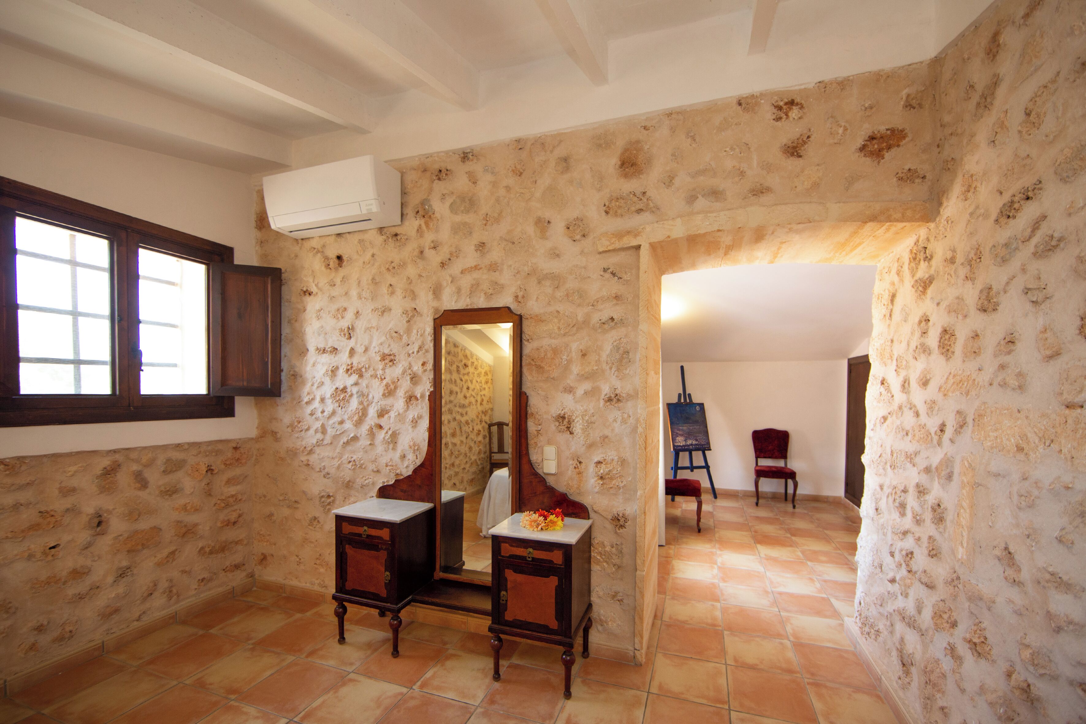 Une pièce aux murs de pierre, avec un meuble en bois, un miroir, une petite table sur laquelle se trouve un vase, et une chaise.
