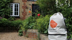 A white bag with "No. 11" and "Number Eleven Great Gransden" text, placed on a brick ledge near a brick building with a window and greenery.