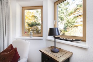 Ein Zimmer mit weißem Stuhl, einem kleinen Holztisch mit Lampe und einem Fenster mit Blick auf Bäume und Gebäude.