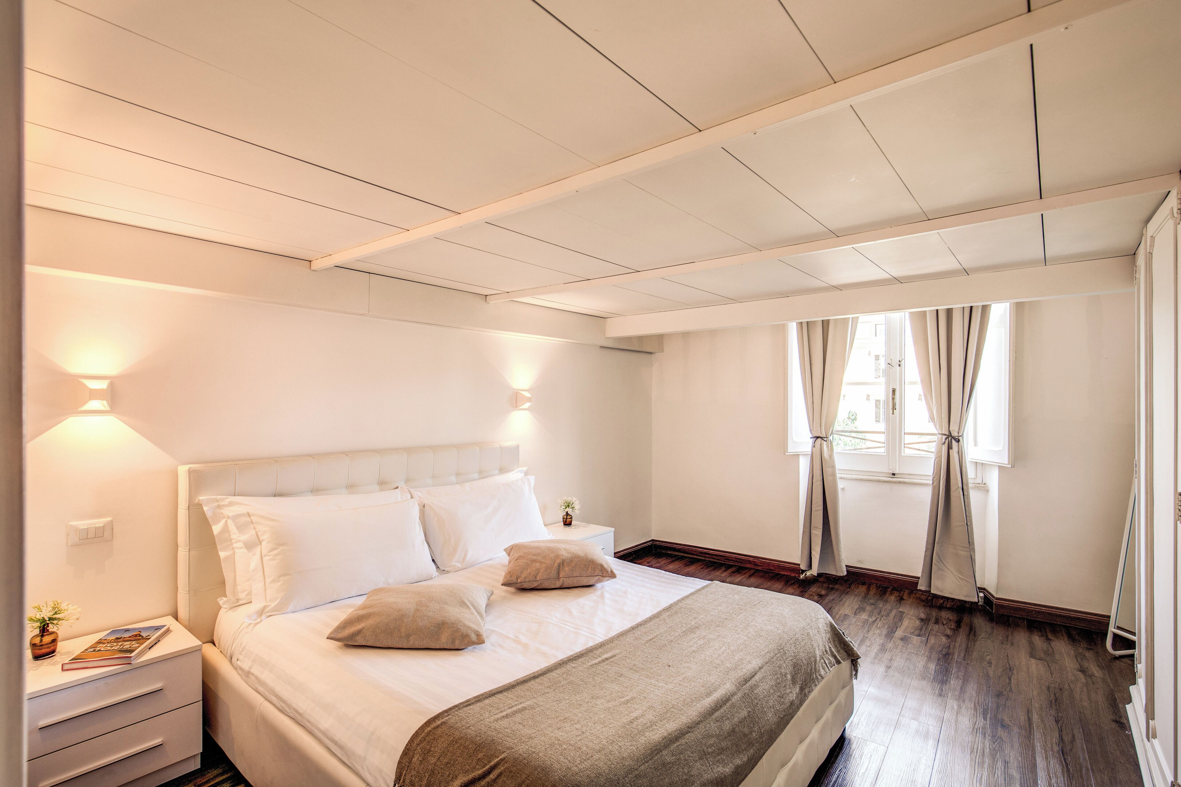 Un dormitorio ordenado con cama, mesitas de noche y una ventana con cortinas.