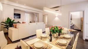 A modern dining area with a long table set for six, surrounded by white chairs. A television is mounted on the wall, and a kitchen is visible in the background.