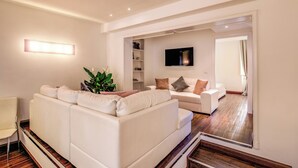 A modern living room with a white sofa, a small table, and a plant. There is a TV mounted on the wall and a door leading to another room.