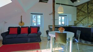 Ein Wohnzimmer mit einem dunklen Sofa, roten Kissen, einem runden Esstisch und einer Küchenzeile mit einer Steinwand und Holzbalken.