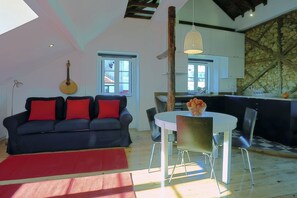 Una sala de estar con un sofá oscuro, cojines rojos, una mesa de comedor redonda y una zona de cocina con pared de piedra y vigas de madera.