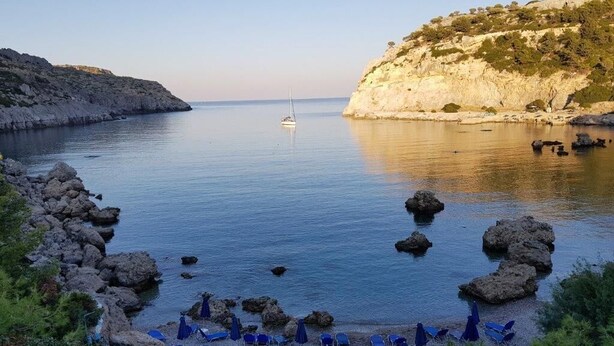 A coastal beach with rocky terrain, a sailboat in the distance, and blue beach chairs and umbrellas. - Brand New Top Luxury Villa!  Stunning modern, extremely stylish. (Afandou)