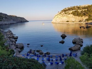 A coastal beach with rocky terrain, a sailboat in the distance, and blue beach chairs and umbrellas.
