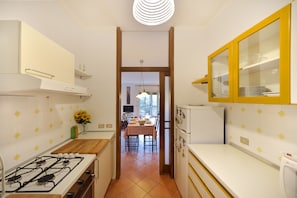 Una cocina con estufa a gas, armarios blancos, un aparador con marco amarillo y un comedor con mantel a cuadros.