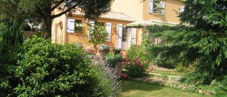 A yellow house with a garden, a large tree, and a stone wall.
