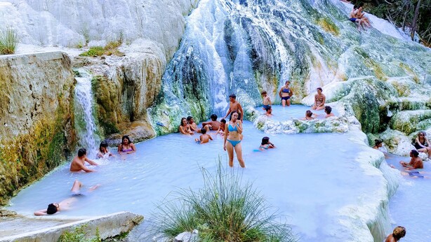 A group of people relaxing in a natural hot spring surrounded by rocky terrain and waterfalls.