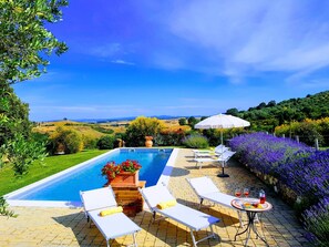 Un espace piscine agrémenté de chaises longues et d’une table, entouré d’un jardin où fleurissent des plantes aux couleurs violettes, et offrant une vue sur des collines ondoyantes.