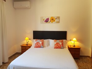 A hotel room with a bed, two bedside tables with lamps, a wall-mounted air conditioner, and a floral painting above the bed.
