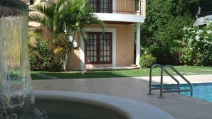 A swimming pool with a water feature in the foreground, a poolside area, and a building with a balcony in the background.