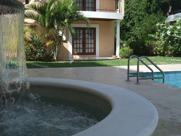 A swimming pool with a water feature in the foreground, a poolside area, and a building with a balcony in the background.