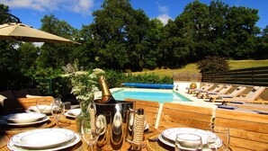Disposición para comer junto a la piscina, con una mesa preparada para cuatro personas, una botella de champán y sillones de descanso al borde del agua.