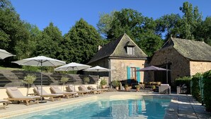 Una piscina con sillas de descanso y sombrillas, rodeada de edificios de piedra y árboles.