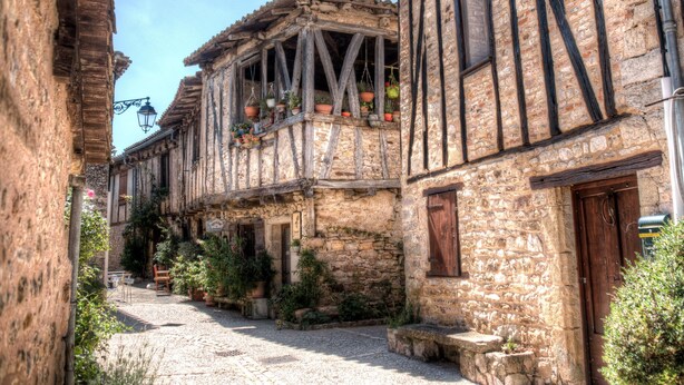 Una estrecha calle empedrada flanqueada por edificios de piedra con entramado de madera y plantas en macetas.