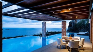 Piscina infinita con vista al mar, pérgola de madera y zona de estar al aire libre.
