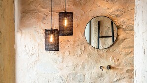 Un baño con pared de piedra, un espejo redondo, un lavabo negro y lámparas colgantes.