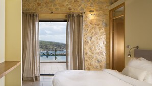 Un dormitorio con pared de piedra, cama, mesita de noche, ventana con cortinas y vista al mar.