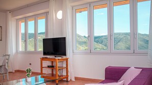 A living room with a purple sofa, a glass coffee table, a TV on a wooden stand, and large windows with a view of hills.