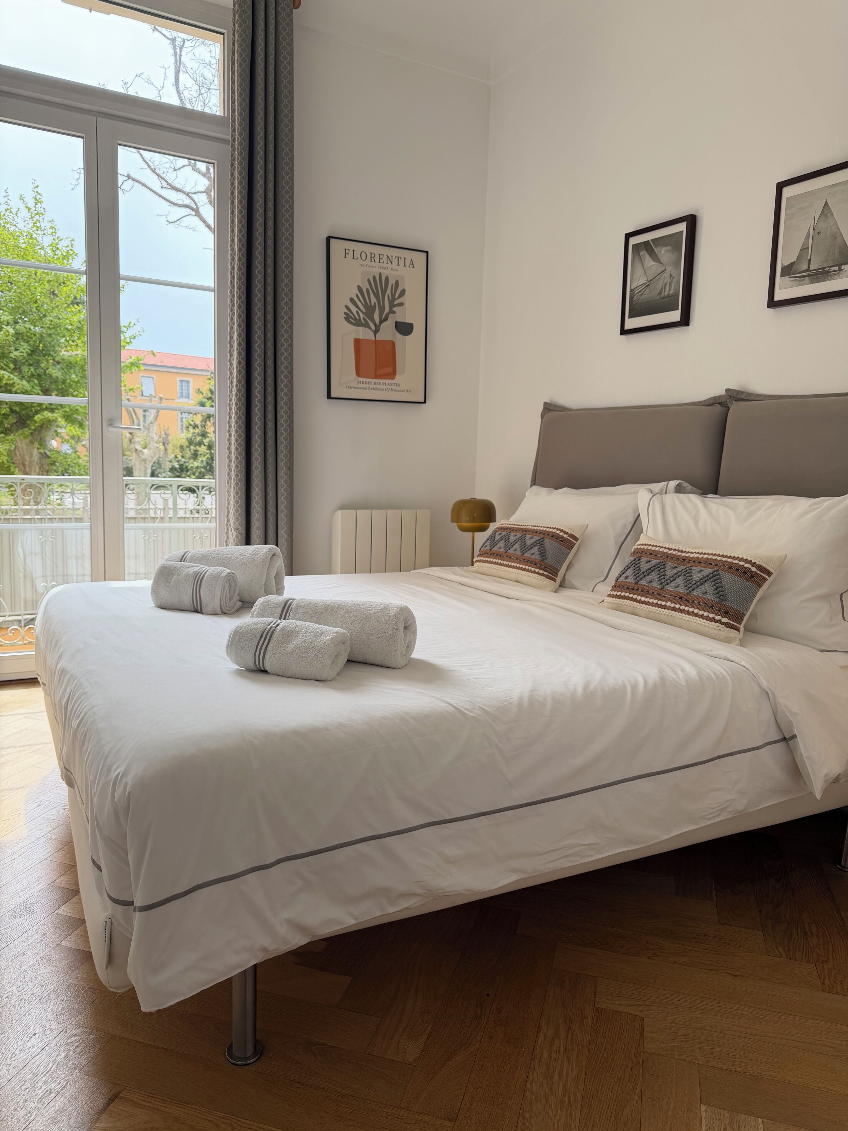 Un lit bien fait, avec des serviettes pliées, un radiateur et des œuvres d’art encadrées accrochées au mur.