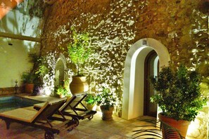 Cortile con pareti in pietra, piscina, poltrone prendisole e piante in vaso.